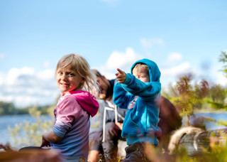 Två glada barn, en pojke och en flicka, sitter runt en mysig lägereld vid vattnet på Orsa Grönklitt - en magisk stund i naturen.