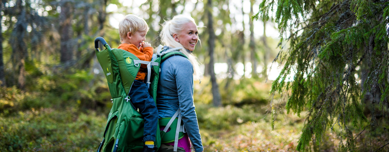 A mother and her son are enjoying a wonderful walk in the forest, with the son sitting in a backpack carrier on his mother's back.