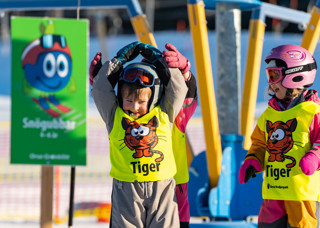 Glada skidskolebarn i slalomhjälm som står framför en liftkarusell i Orsa Grönklitt