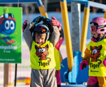 Glada skidskolebarn i slalomhjälm som står framför en liftkarusell i Orsa Grönklitt