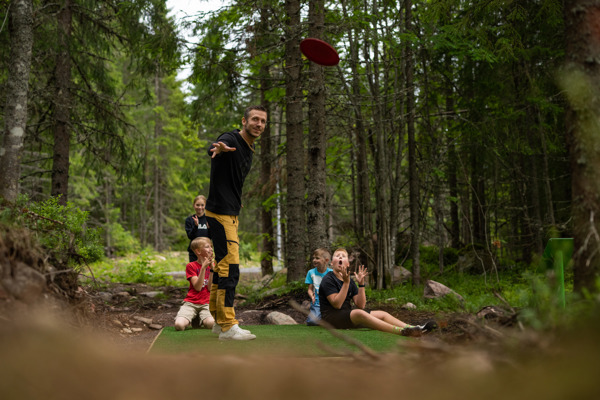 En familj spelar discgolf i Orsa Grönklitt - pappan kastar discen med glädje, medan barnen hejar på i den vackra naturen.