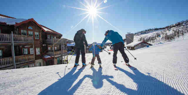 Två föräldrar håller ett barn i handen och åker skidor tillsammans ner för backen